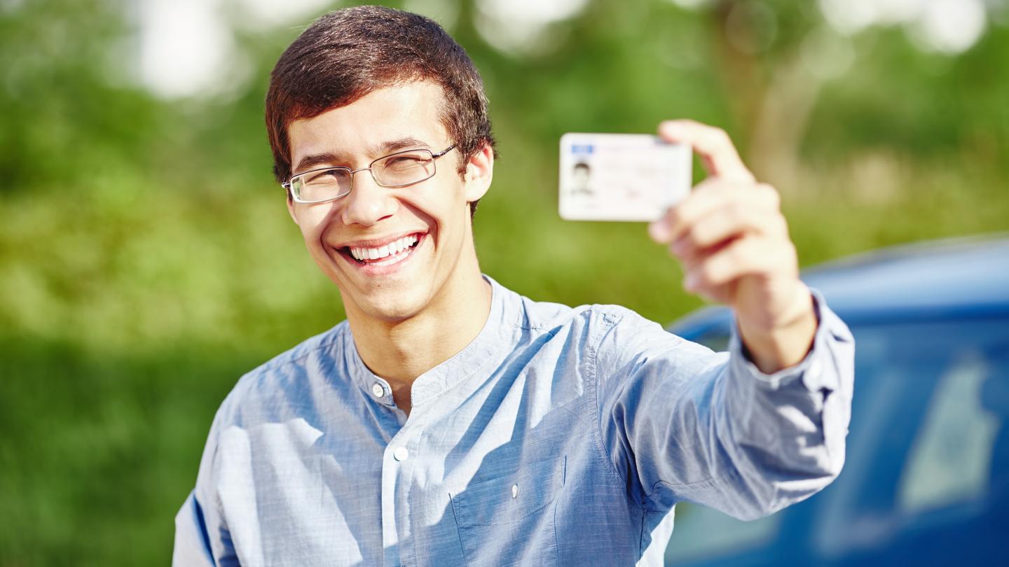 young man in blue shirt and glasses smiling outside holding up driver's license